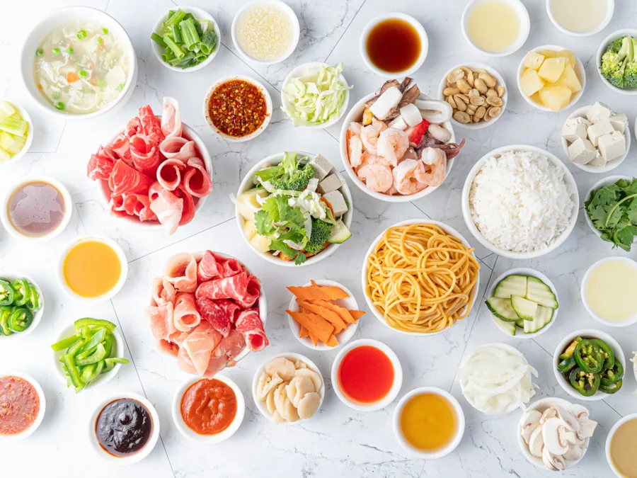 Overhead spread of Mongolian BBQ bar ingredients: sliced beef and pork, shrimp, tofu, rice, noodles, vegetables, peanuts and house sauces